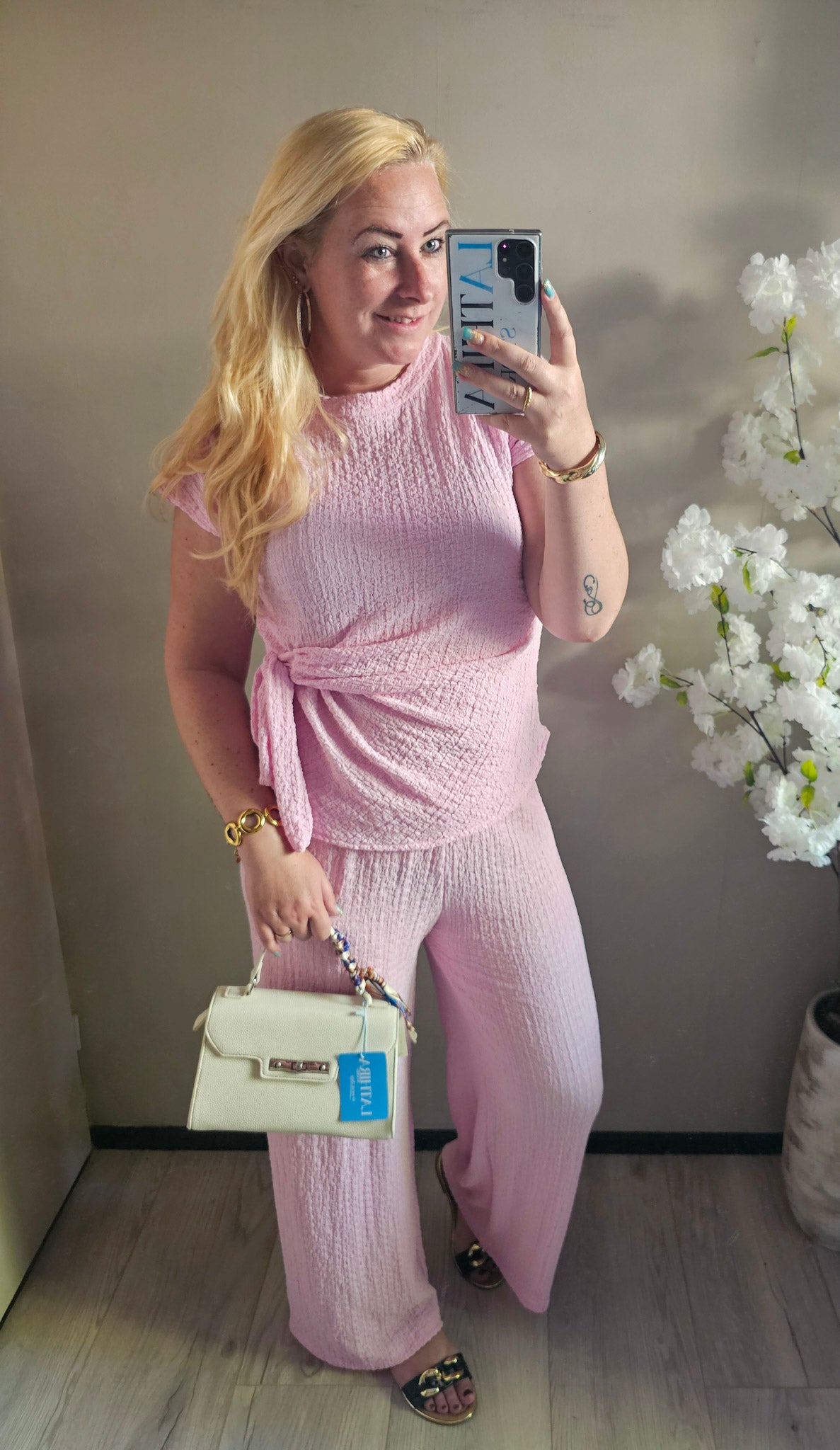 Damesmodel draagt een roze tweedelige set met overslagtop, strikdetail en wijde broek van structuurstof.
