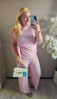 Damesmodel draagt een roze tweedelige set met overslagtop, strikdetail en wijde broek van structuurstof.